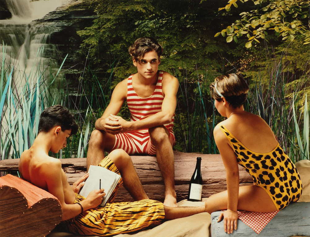 Three young people in bathing suits lounging on a constructed beach with a waterfall and forest in the back.